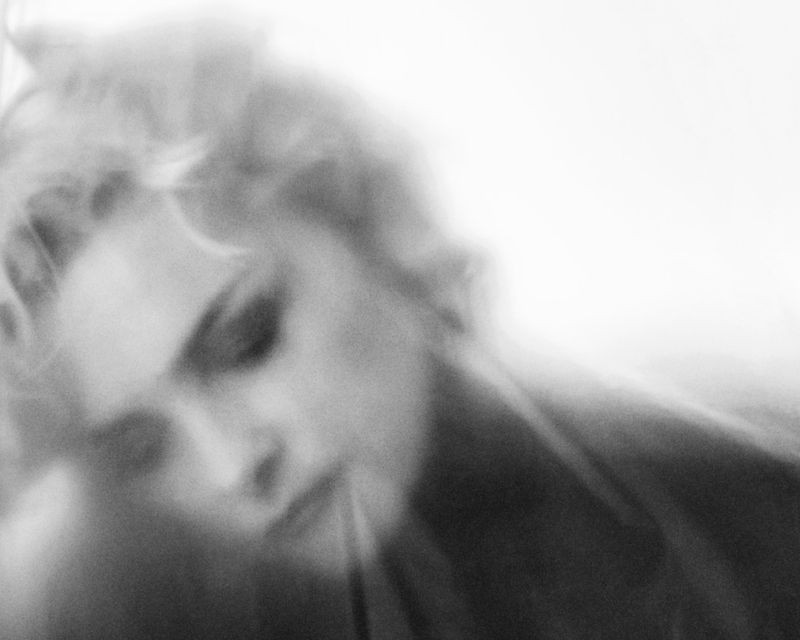 Monochrome portrait of a blonde-haired woman with diffused focus, downward gaze, and high-key lighting in a photography style.