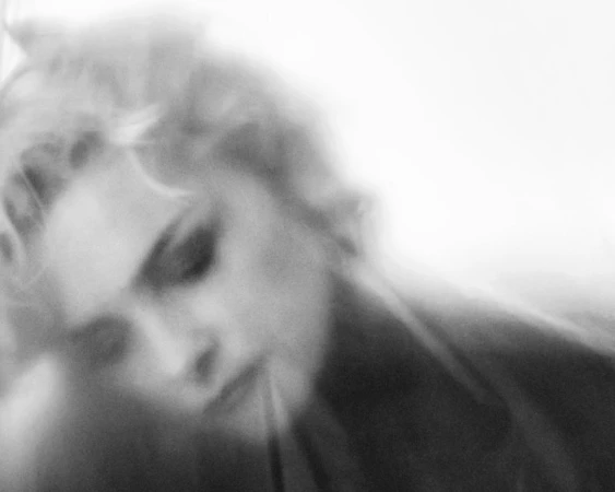 Monochrome portrait of a blonde-haired woman with diffused focus, downward gaze, and high-key lighting in a photography style.
