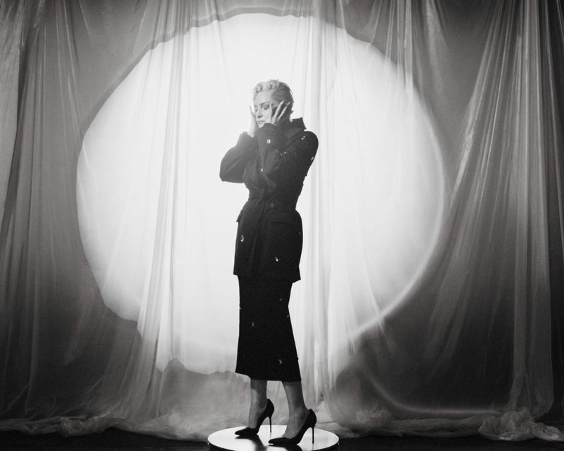 Black and white portrait of a fair-skinned adult woman with short blonde hair posing in high heels against a bright backdrop.