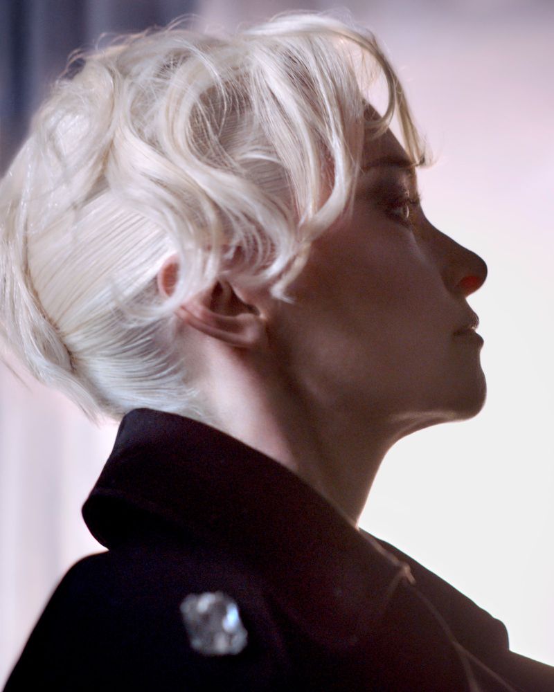 Headshot of a woman with short platinum blond hair wearing a dark coat with a jeweled brooch. Her face is in profile view.