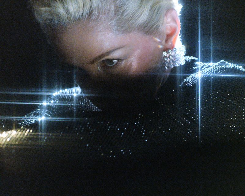 Headshot of an adult woman with short blonde hair wearing sparkly jewelry and a black dress, captured in a low-key, studio setting.