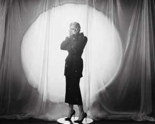 Alison Goldfrapp posing in a black embellished jacket and heels in front of a circular backlit sheer curtain.