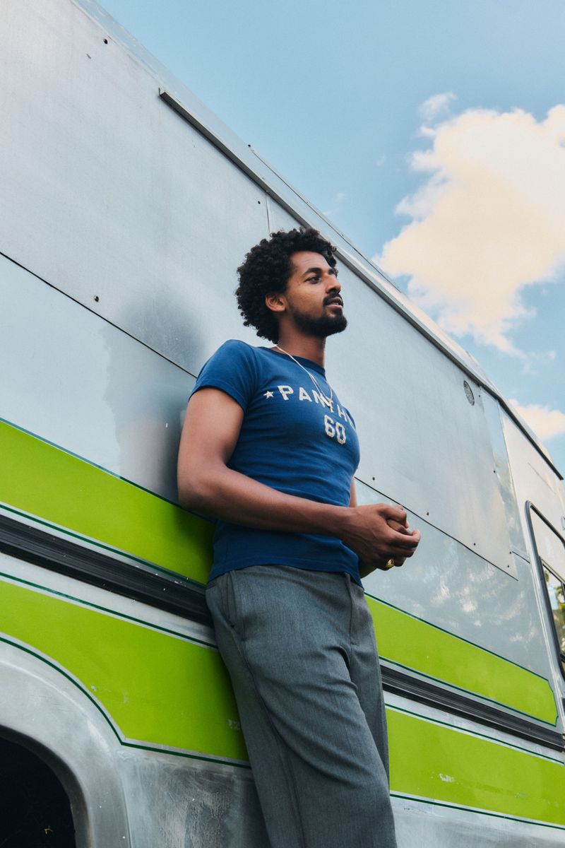 Adult male with curly hair leans against a silver vehicle with a green stripe under a blue sky with wispy clouds. He wears a blue t-shirt.