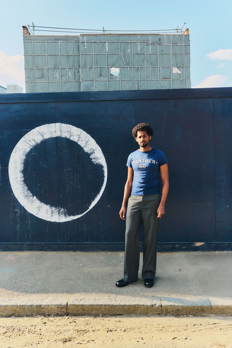 Standing man in casual t-shirt and trousers poses against a black wall with a white circle design, with an urban backdrop and blue sky.