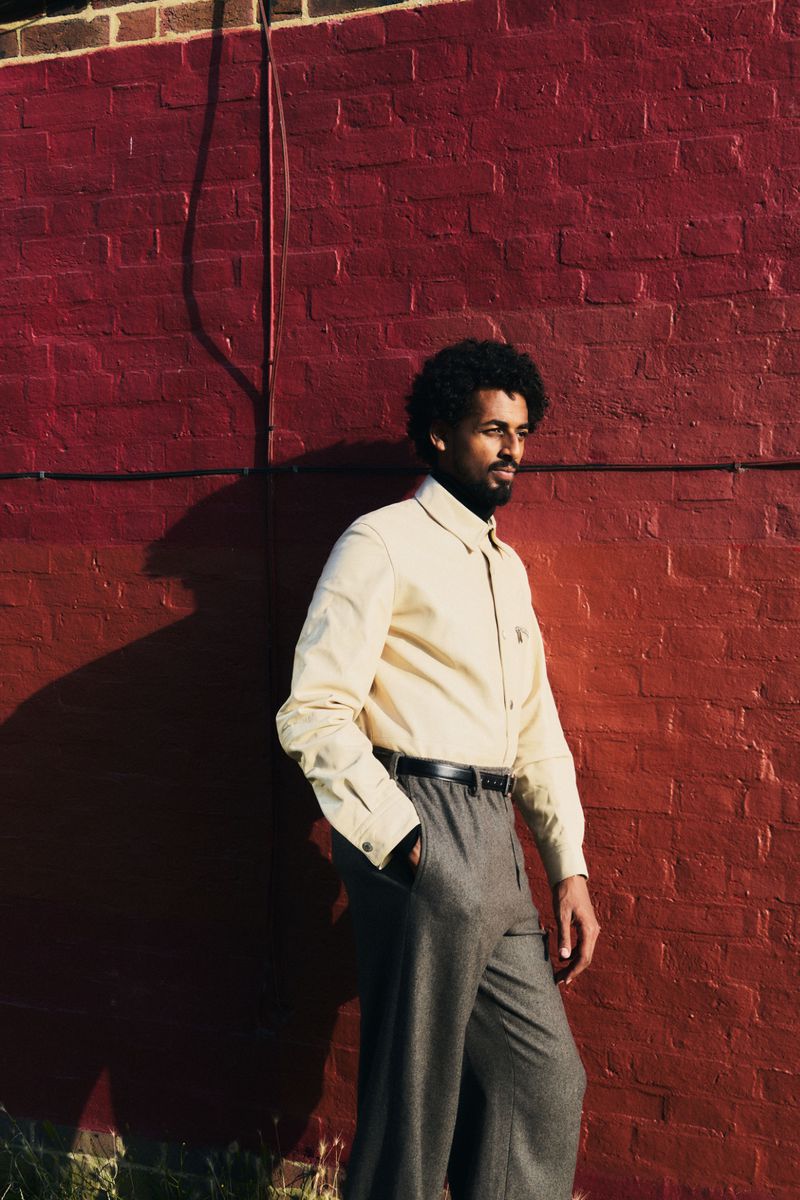 Michael Workeye standing in a cream button-down shirt and grey pleated trousers against a painted red brick wall in direct sunlight.