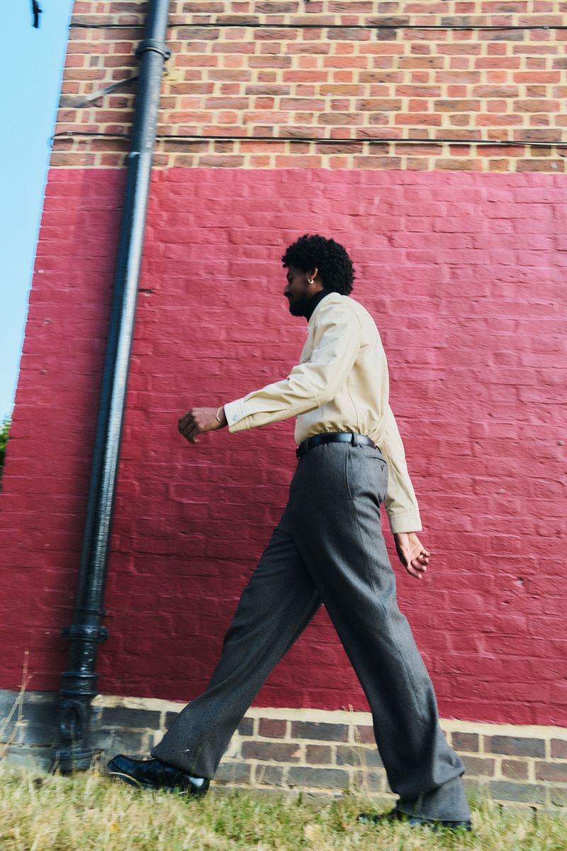 A man in a tan shirt and grey pants walks outdoors on the grass next to a brick wall. He is walking away from the camera.