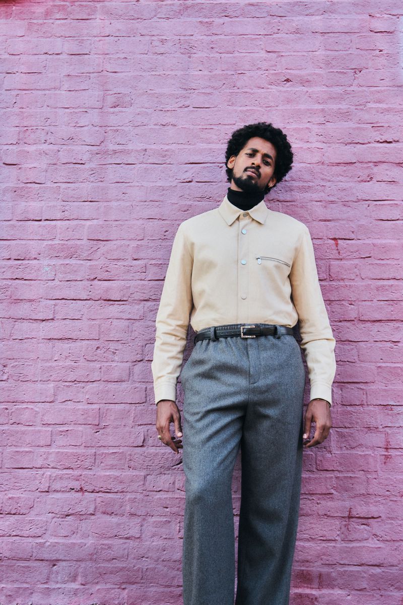 Adult male with beard and curly hair, wearing button-down shirt and grey trousers, posing in front of a textured pink brick wall.