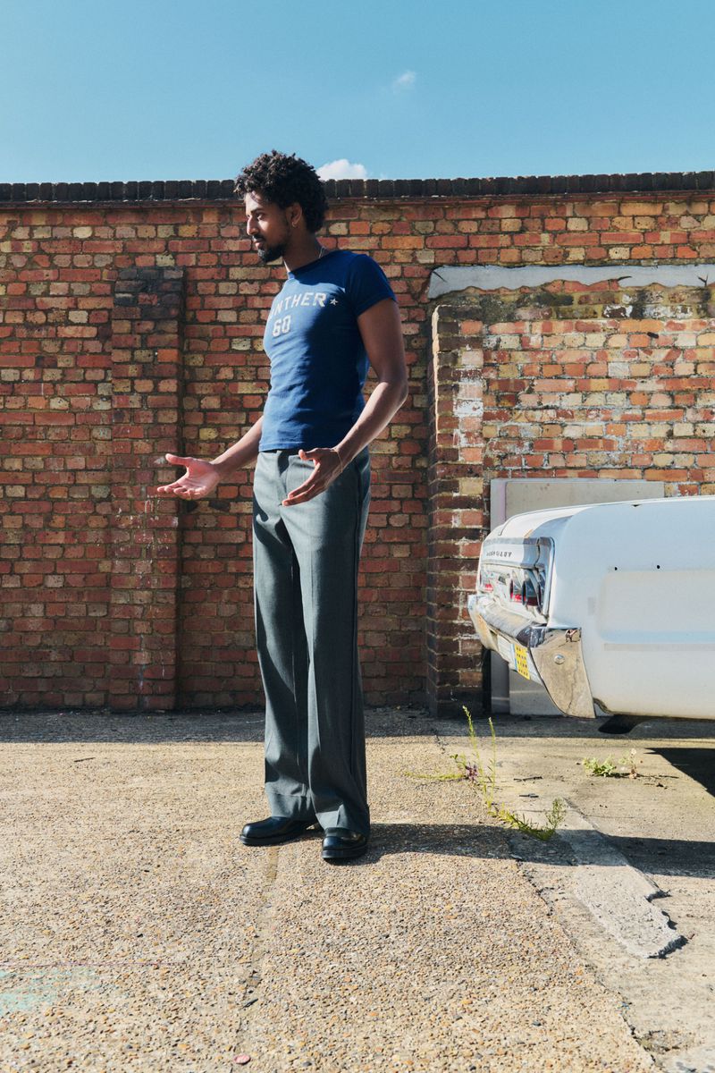 Adult man in blue t-shirt and grey pants poses on cracked concrete with a white car and brick wall in the background on a sunny day.