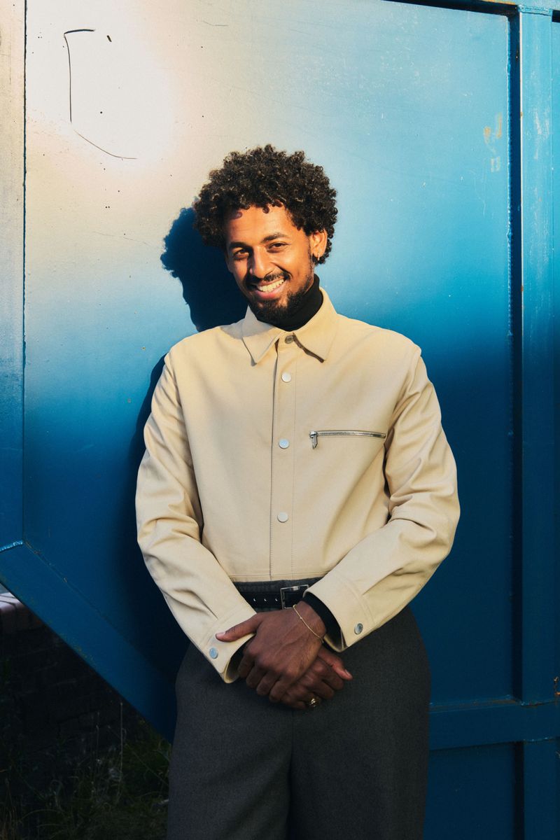 Portrait of a smiling, dark-haired male leaning against a blue wall, wearing a beige shirt, black turtleneck, and gray pants outdoors.