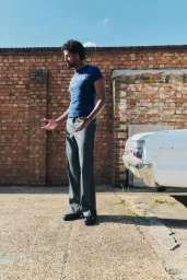 Michael Workeye standing outdoors in a blue graphic t-shirt and wide-leg trousers in front of a brick wall and vintage white car.