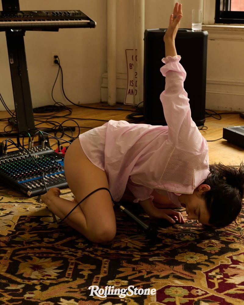 Woman in pink top kneels on patterned rug with music equipment; microphone is centered in foreground; Rolling Stone logo visible.