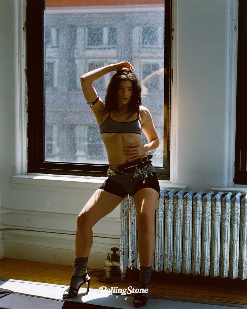 Singer/songwriter poses in front of a window with city views in a studio-lit interior. She wears a sports bra, shorts, and heeled shoes.
