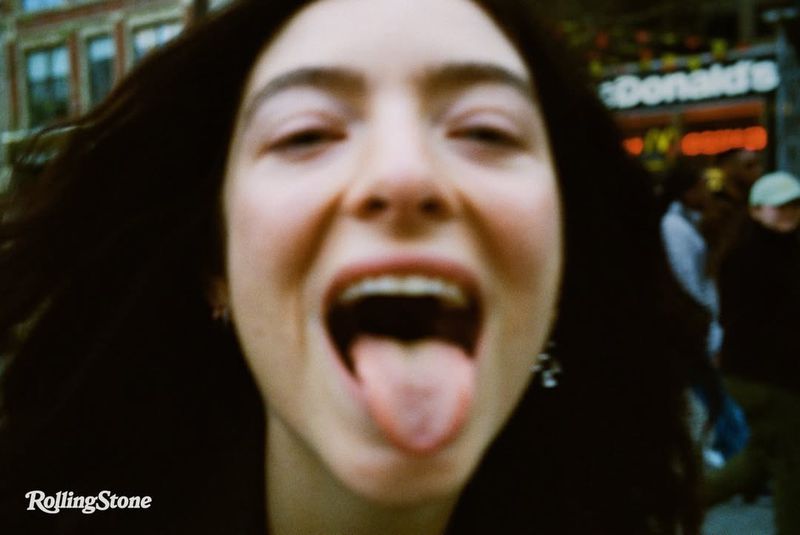 Close-up of a woman sticking her tongue out, with buildings and a restaurant visible in the blurred background.