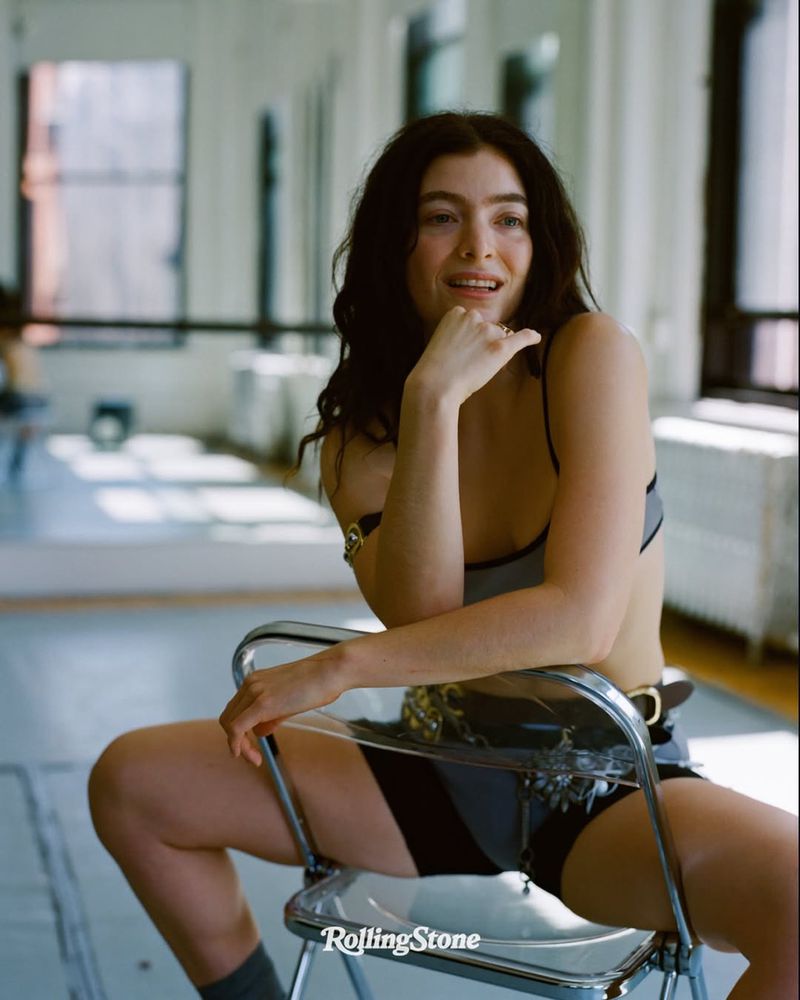 Woman sits in a transparent chair in a photo studio. She wears a bra and shorts and is posed with her hand near her face.
