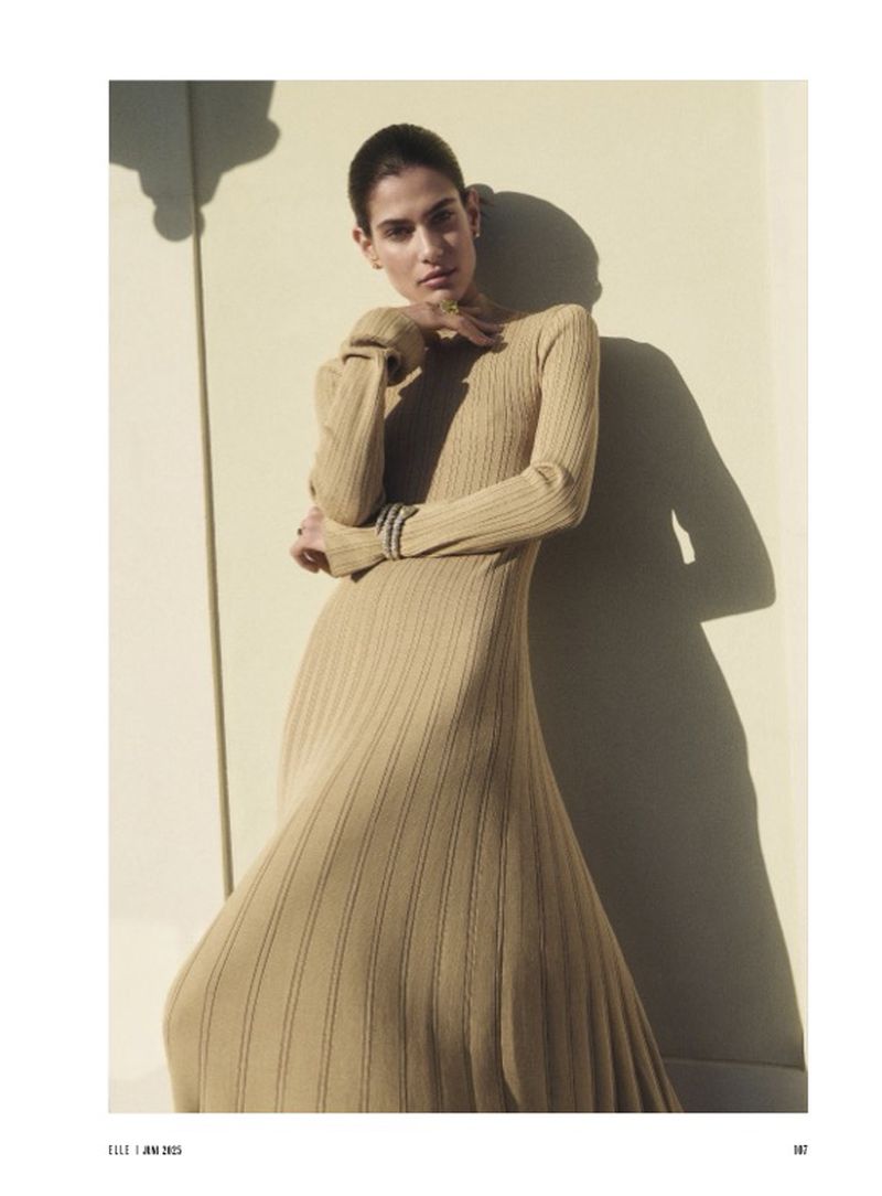 Indoor portrait shot of a woman with short dark hair in a tan ribbed, long-sleeved dress posing next to a wall with soft lighting.