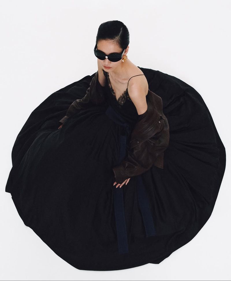 Woman in a black gown and sunglasses, with a brown leather jacket, poses against a white background in a fashion editorial shot.