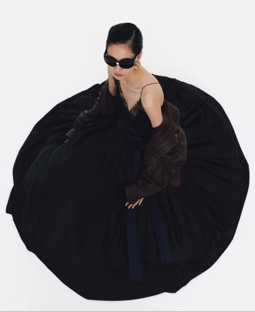 Woman in a black gown and sunglasses, with a brown leather jacket, poses against a white background in a fashion editorial shot.