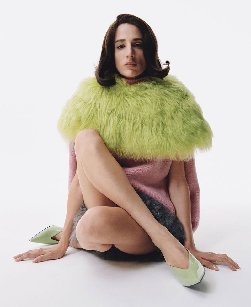 Woman poses seated on a white seamless background wearing a pink sweater, bright-green faux fur collar, and matching green pointy shoes.