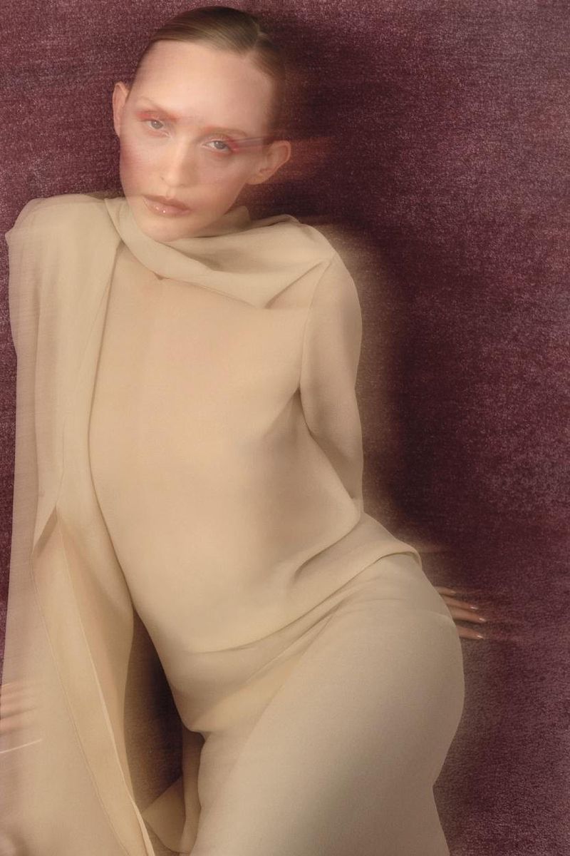 Elegant portrait of an adult woman in a flowing beige dress against a textured purple background. Soft lighting and blurred motion.