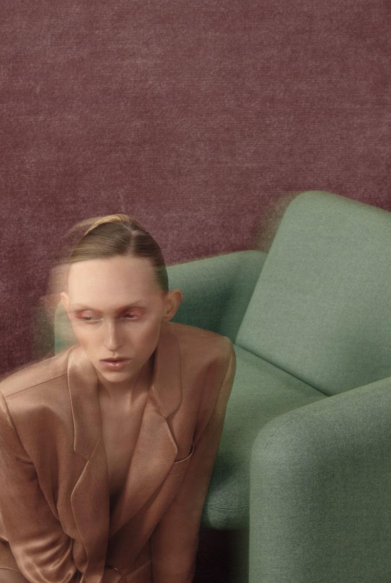 Woman in a silky peach blazer sits on a light green couch against a textured plum backdrop in a fashion portrait with soft focus.