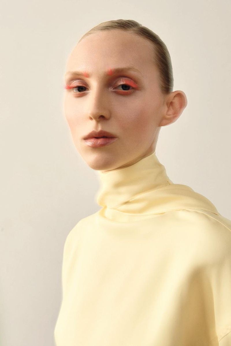 Close-up studio portrait of a fair-skinned young woman with bright orange eye makeup, wearing a pale yellow turtleneck top.
