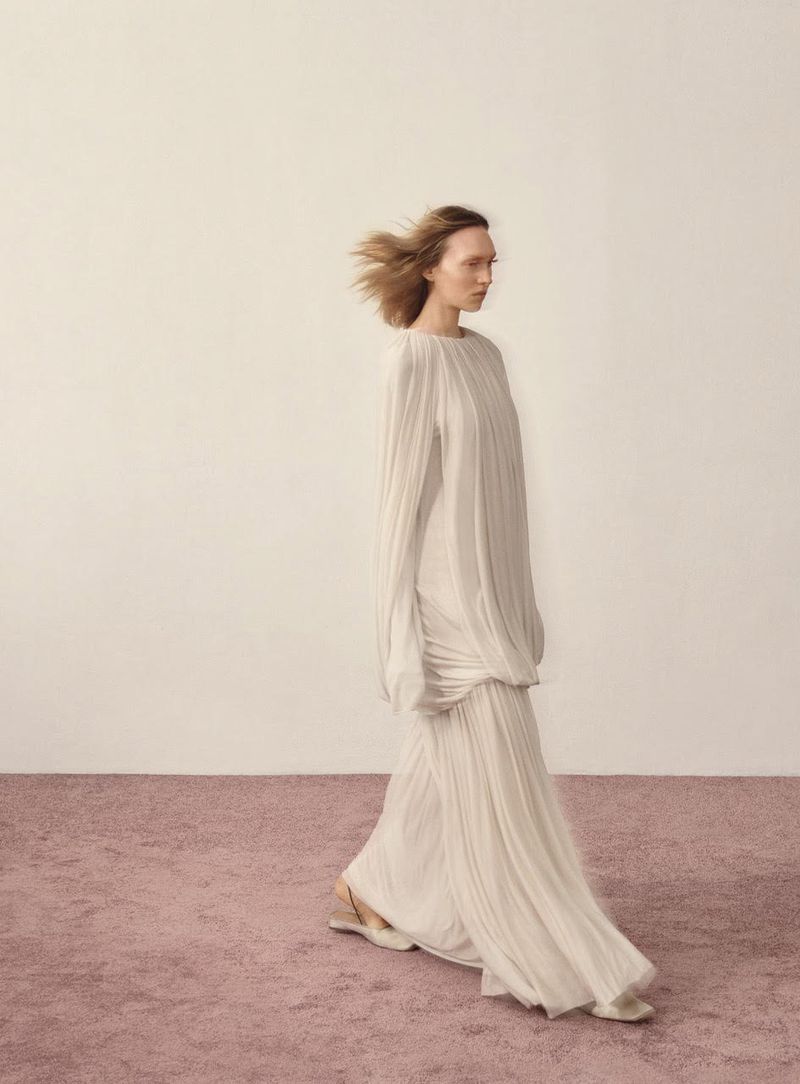 Woman in flowing white dress walks against a neutral backdrop with a textured pink floor, creating an elegant and minimalist fashion image.