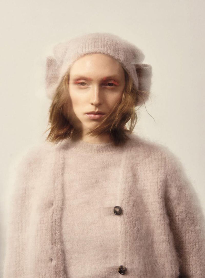 Close-up portrait of a woman wearing a fuzzy pink hat and cardigan sweater in a photo studio setting with soft lighting and light color grading.