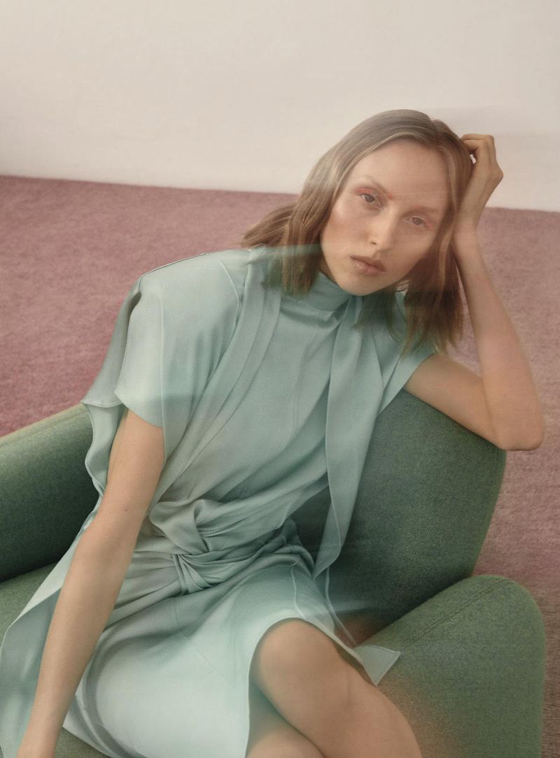 A fair-skinned woman with wavy blonde hair sits in a green chair, wearing a light green midi dress in a studio with a pink carpet.