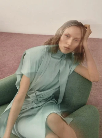 A fair-skinned woman with wavy blonde hair sits in a green chair, wearing a light green midi dress in a studio with a pink carpet.