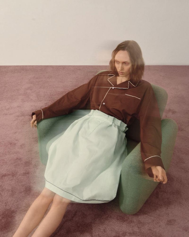 A woman with auburn hair sits in a green chair in a brown shirt and light blue skirt, posed in a photo studio with a white and mauve wall.