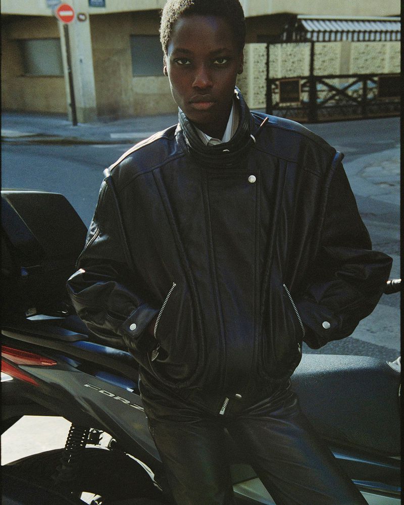 A young Black woman models a black leather jacket and pants while leaning against a black scooter on a city street.