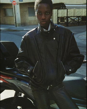 A young Black woman models a black leather jacket and pants while leaning against a black scooter on a city street.