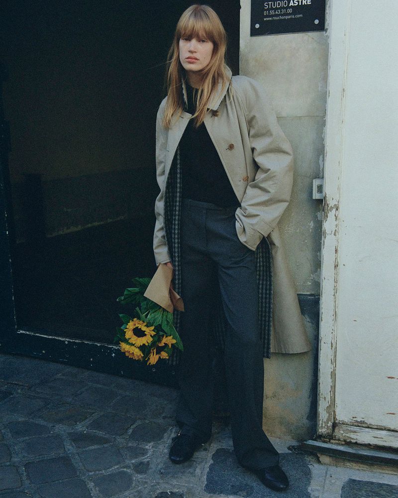 Woman in trench coat holds sunflowers standing near a doorway with stone walls, wearing black top and grey pants, in an urban setting.