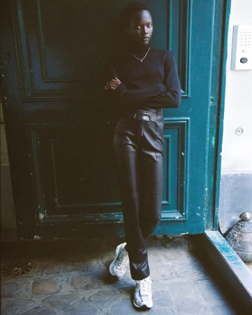 A South Sudanese adult male stands in front of a blue door wearing black clothes and white sneakers in a medium shot with an eye-level angle.