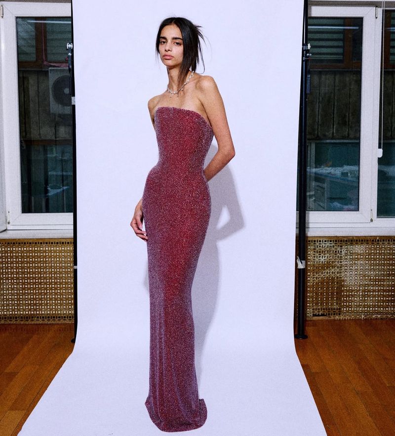 A slim adult female poses in a sparkly red strapless gown against a white backdrop with studio lighting and a window in the background.