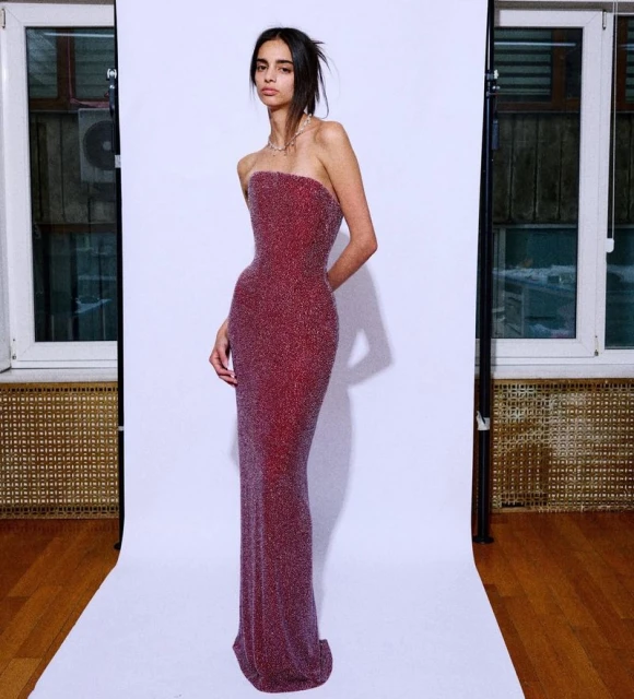 A slim adult female poses in a sparkly red strapless gown against a white backdrop with studio lighting and a window in the background.