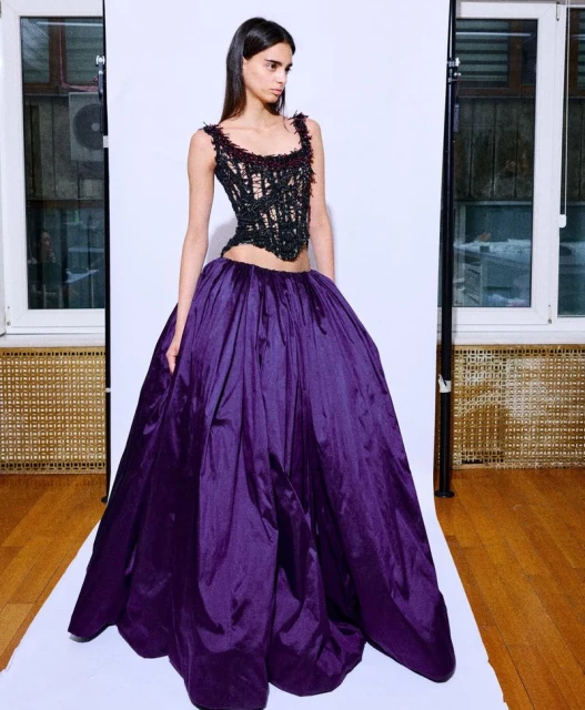 Adult female fashion model poses in a black corset and voluminous purple ball gown in a photo studio against a white backdrop and wooden floor.