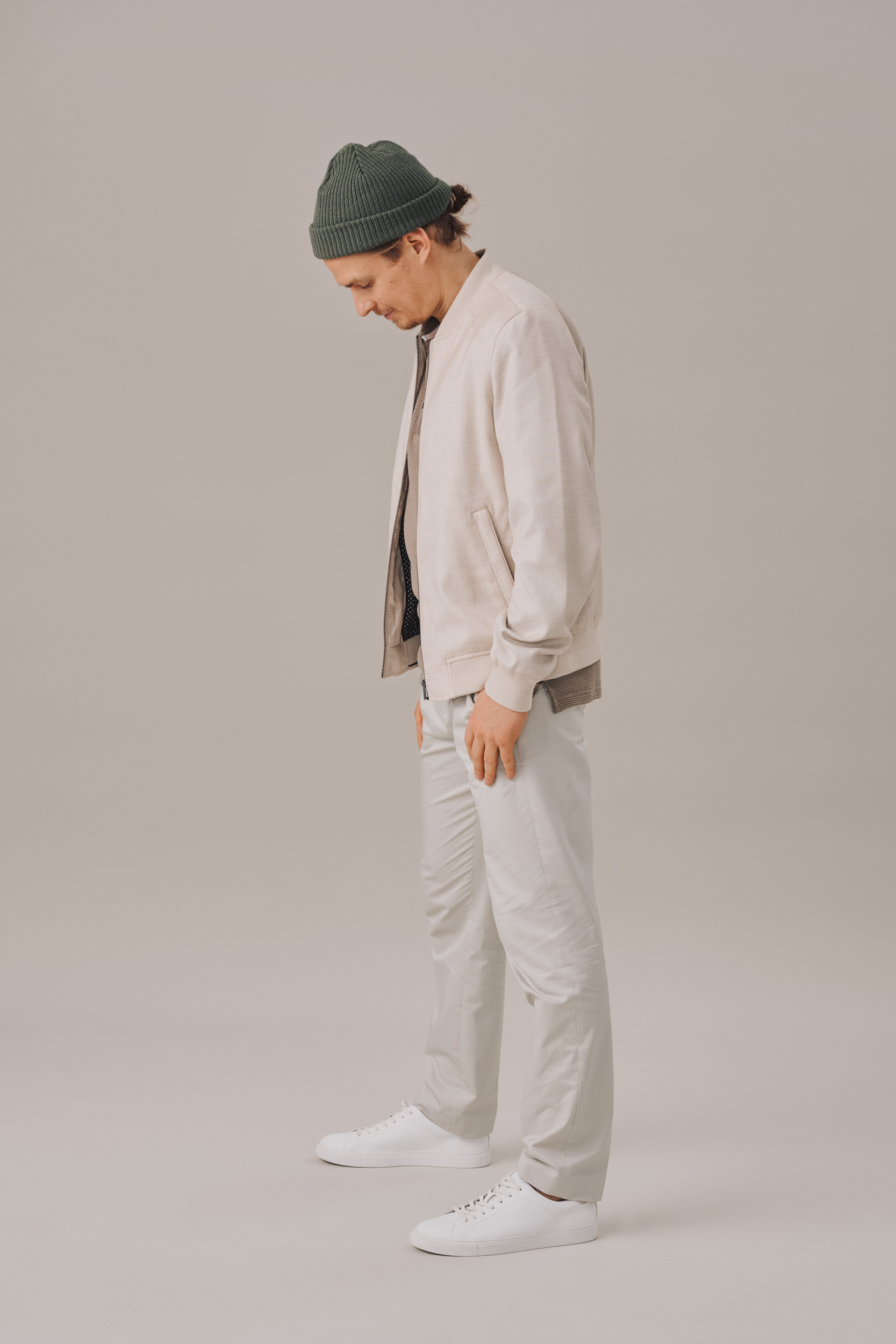 Man in light jacket, grey pants and white sneakers stands with head down in a studio. He wears a dark green beanie.