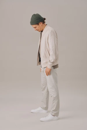 Man in light jacket, grey pants and white sneakers stands with head down in a studio. He wears a dark green beanie.