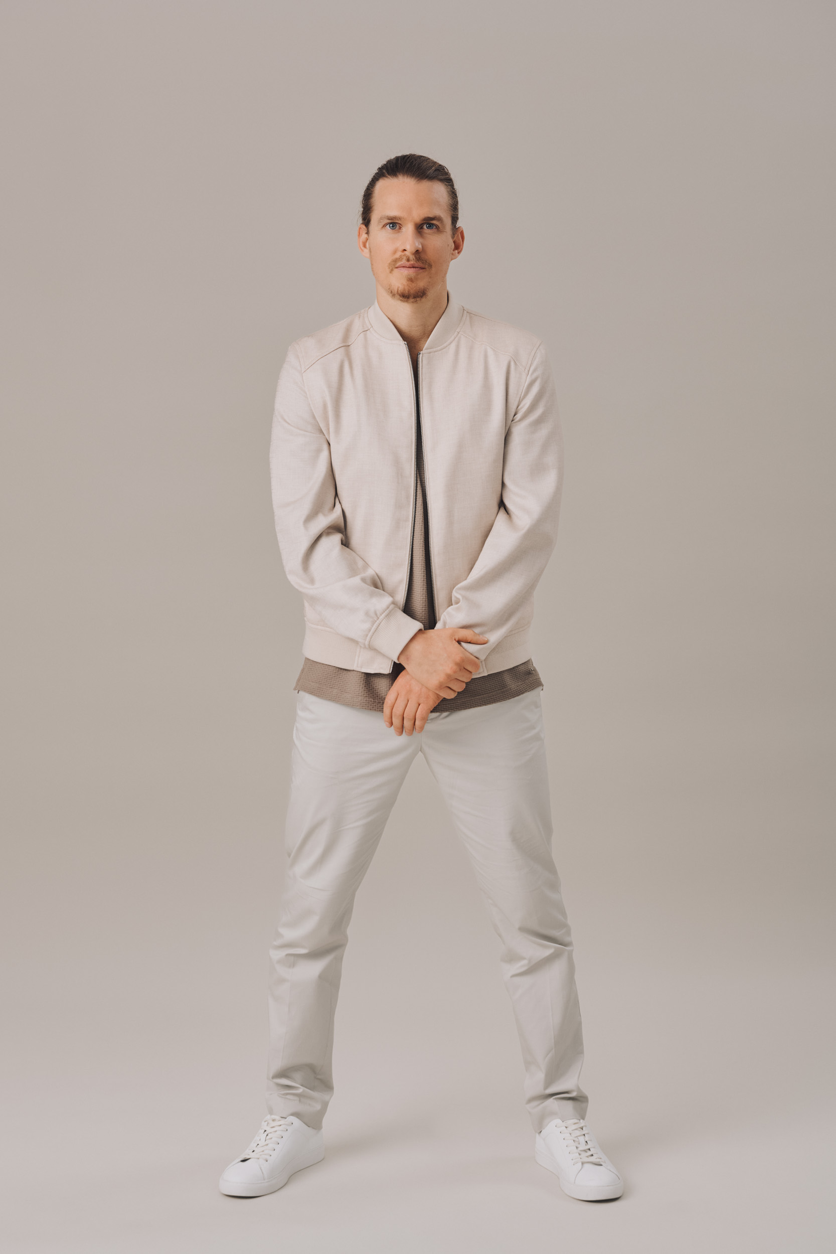 Full-length shot of a Caucasian adult man in casual fashion, wearing a light bomber jacket, light pants and sneakers, posing in a photo studio.