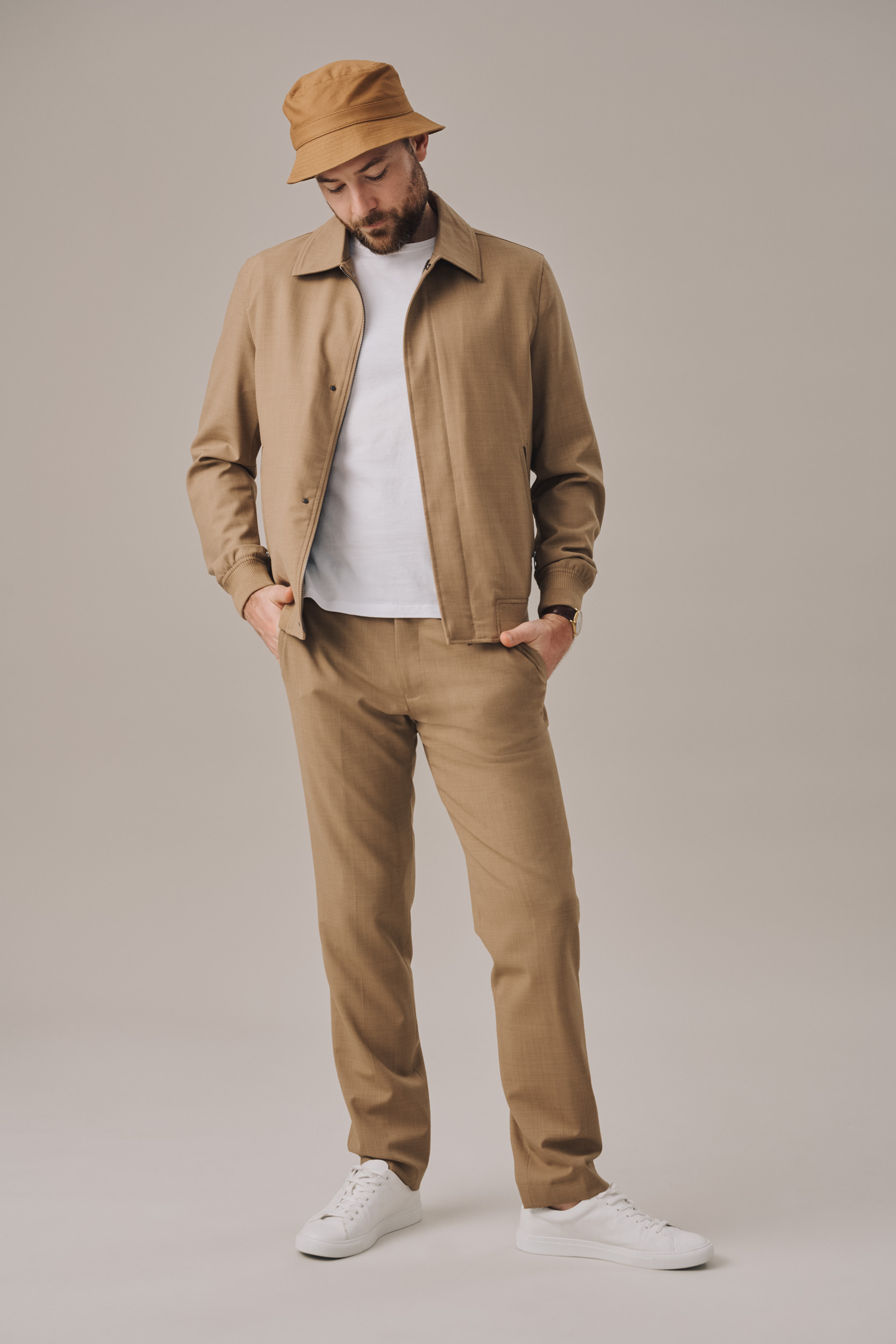 Full shot of an adult male in a matching tan jacket, pants, and bucket hat, standing against a neutral background with white sneakers.