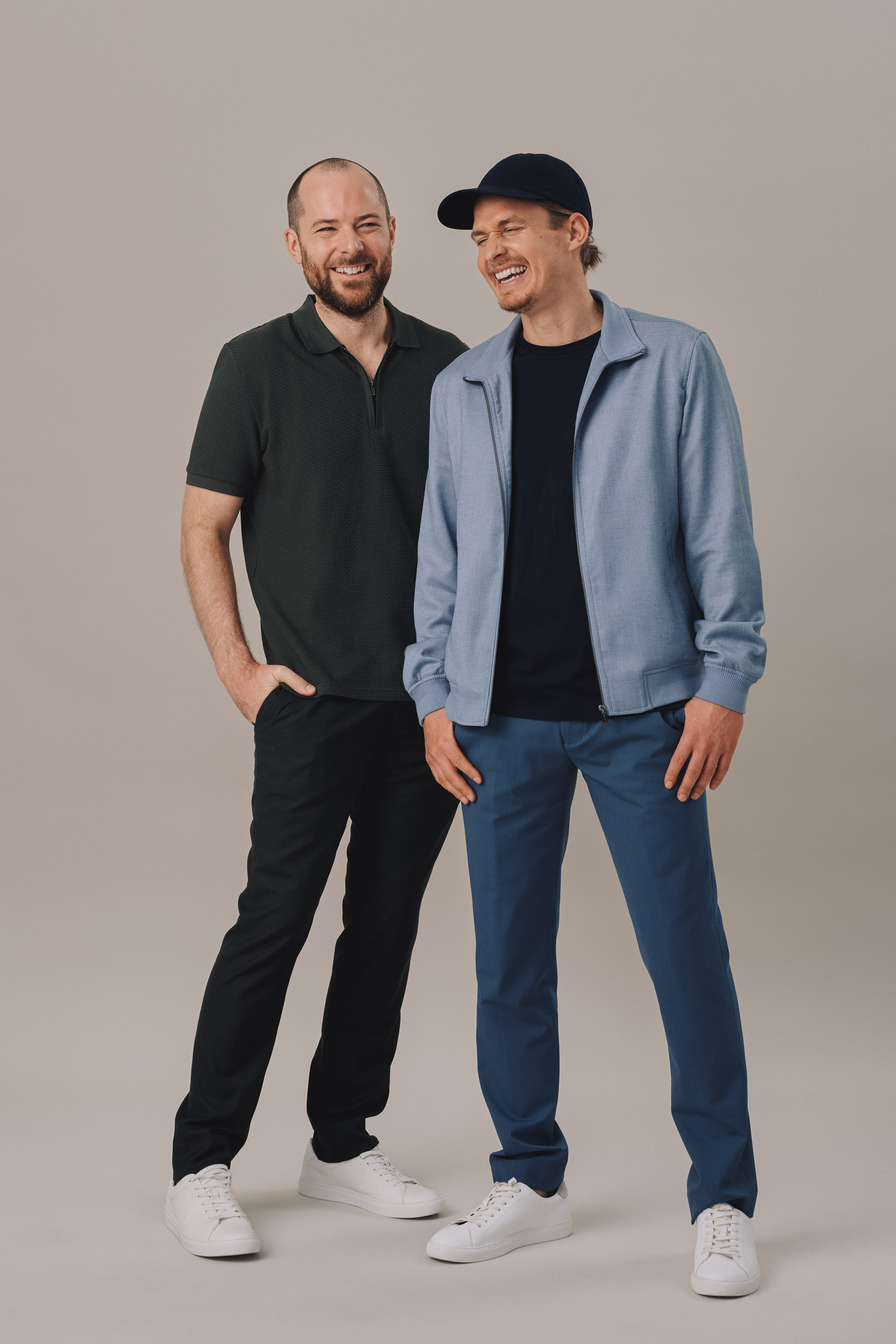 Two adult men stand smiling in casual clothes. One wears a dark green polo and the other a blue jacket and baseball cap.