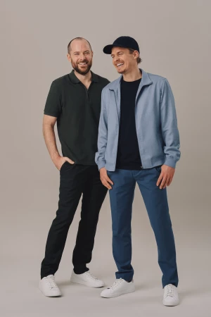 Two adult men stand smiling in casual clothes. One wears a dark green polo and the other a blue jacket and baseball cap.