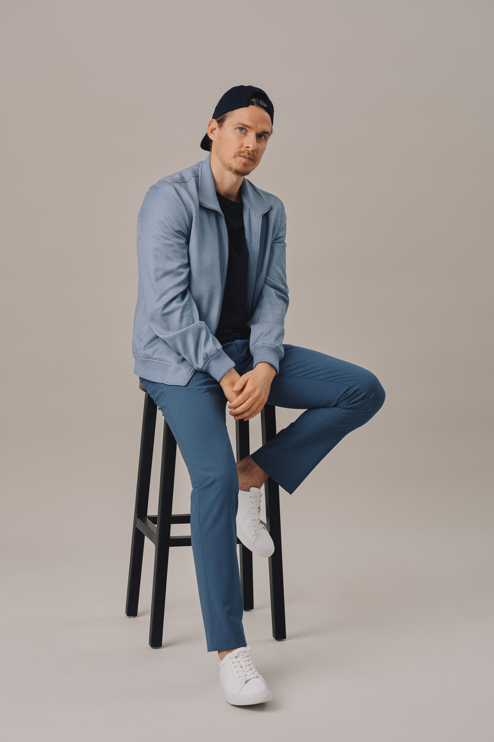 Man in blue jacket and pants sitting on stool. Studio portrait of male model wearing casual fashion, white sneakers and baseball cap.