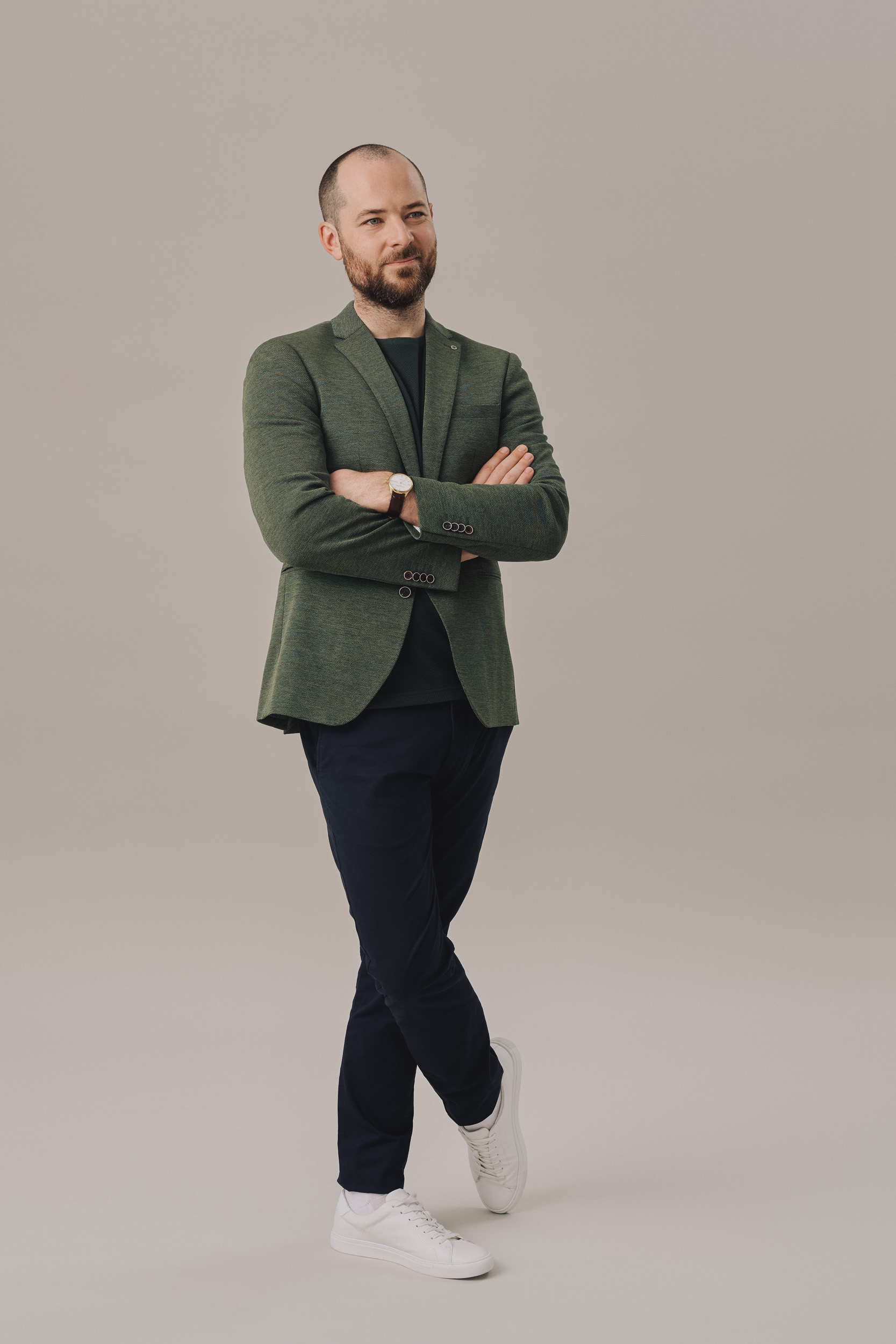 Full shot of a man with a beard in a green blazer and navy pants standing in front of a neutral background with arms crossed.