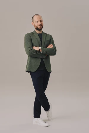 Full shot of a man with a beard in a green blazer and navy pants standing in front of a neutral background with arms crossed.