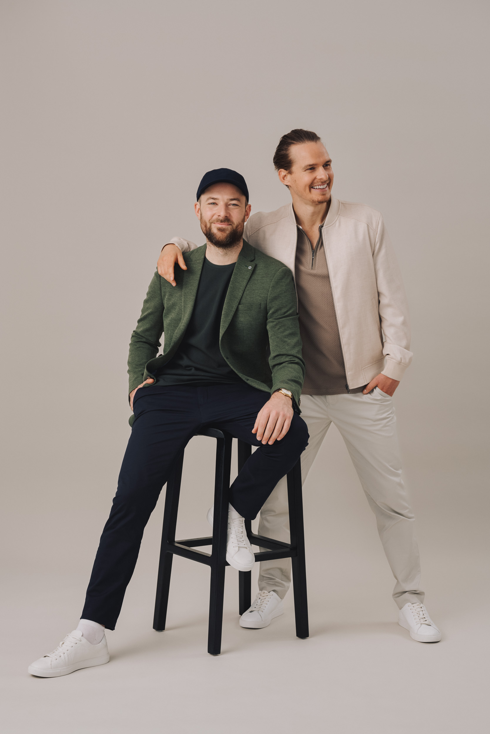Two men, one seated on a black stool, the other standing with arm around the seated man's shoulder, both smiling in studio.