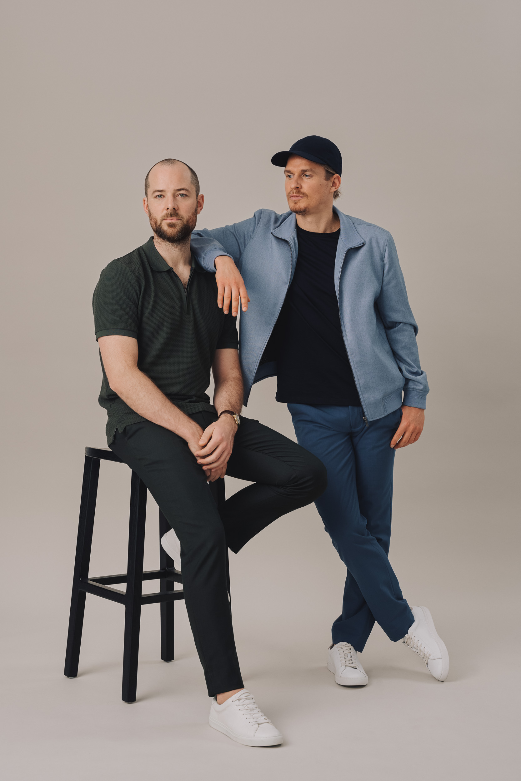 Two adult men in casual attire pose in a studio setting. One man is sitting on a black stool, while the other is standing next to him.