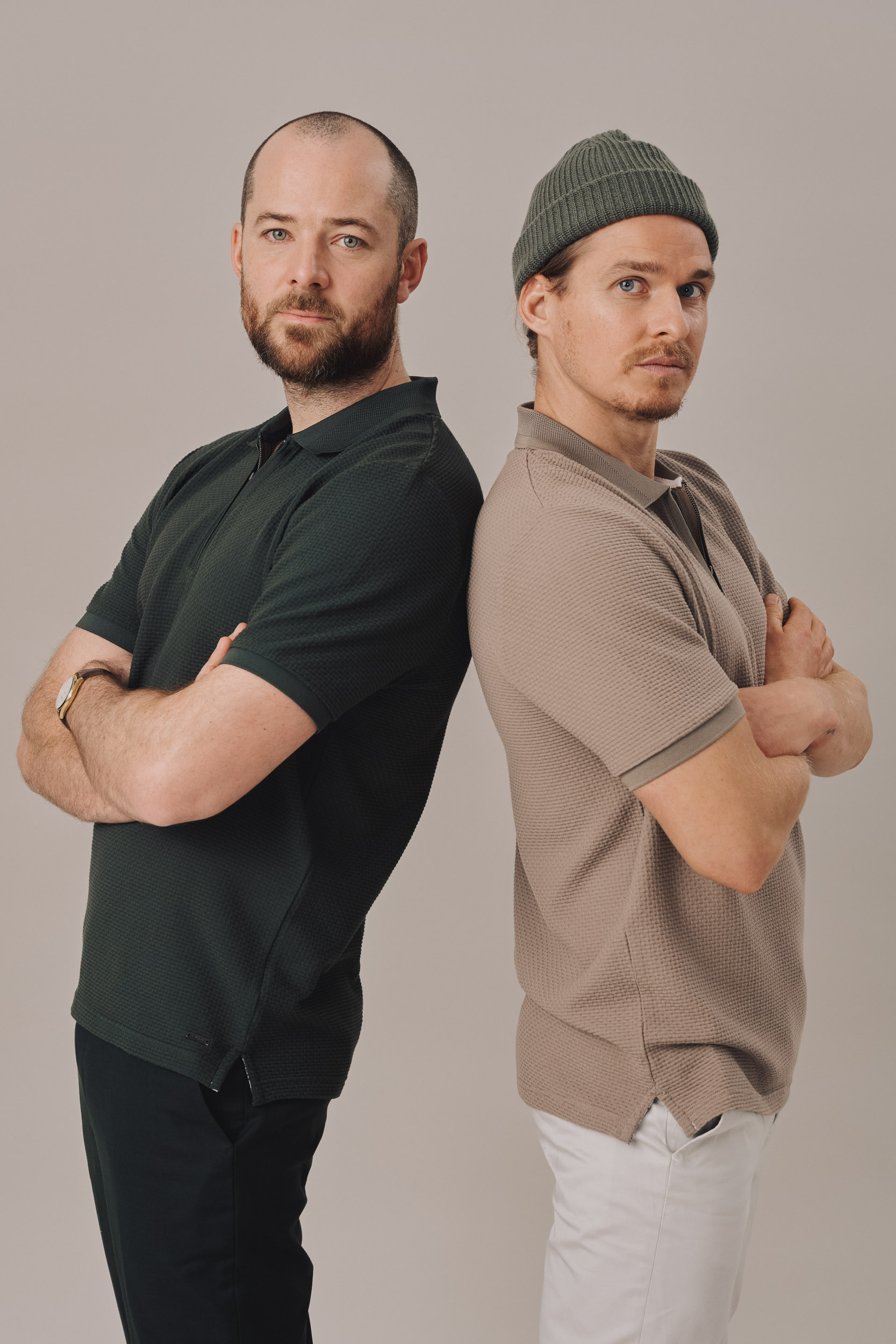 Two adult men pose back-to-back in a studio. One has a beanie, fair skin and a tan polo. The other has dark hair, dark polo and gold watch.