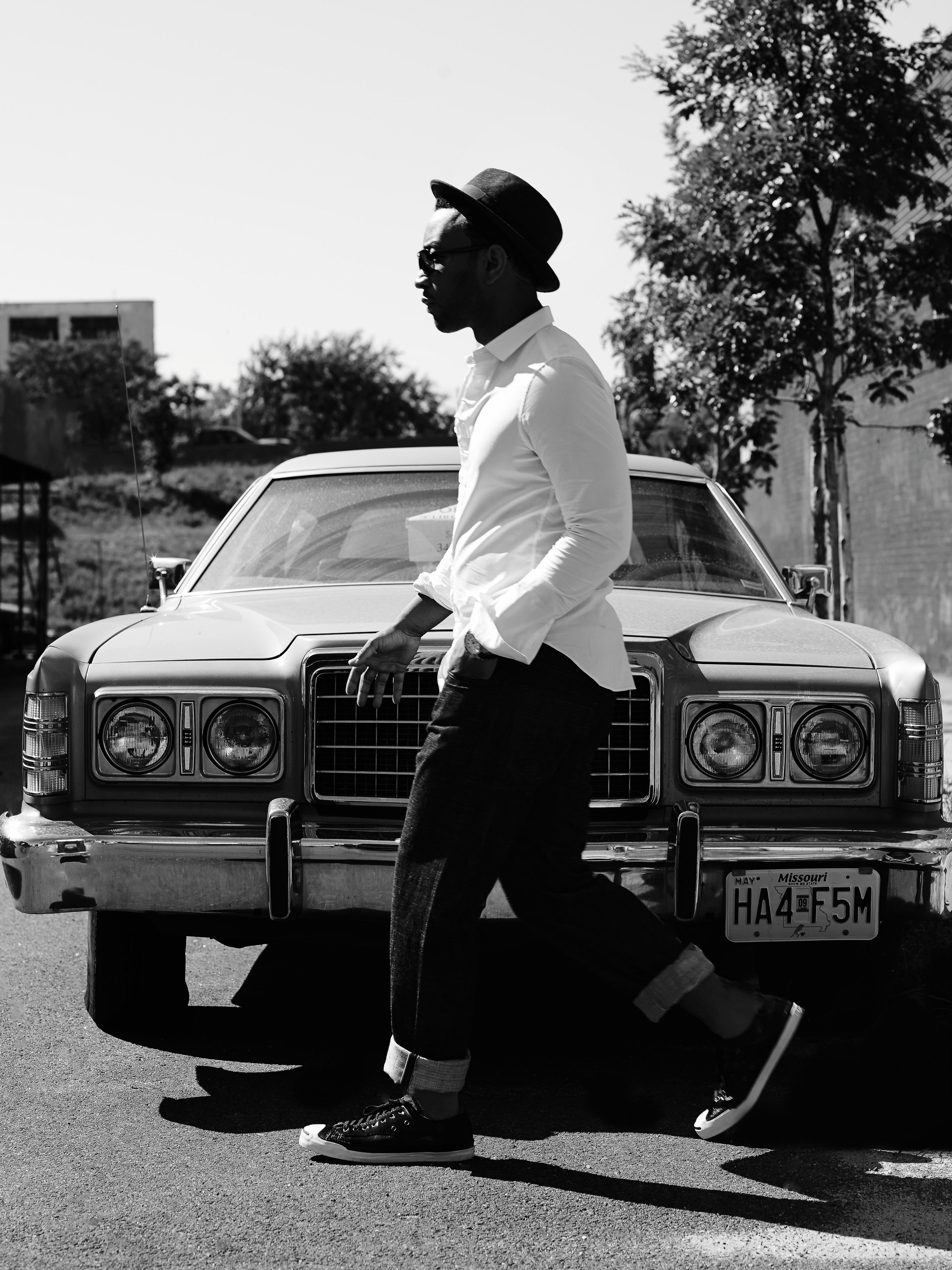 A black and white shot features a man in hat and white shirt walking past a vintage car on a street. The photograph is in black and white.